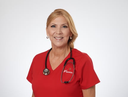 Jenniver Evrard stands smiling in front of a white background in a red BAYADA branded scrub top, blonde hair pulled back and stethoscope around her neck