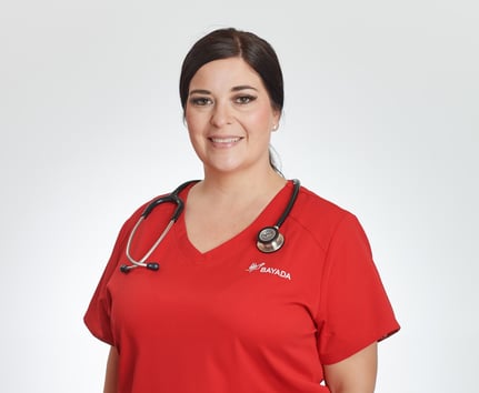 Jennifer Feller stands, smiling, in front of a white background in a red BAYADA branded scrub top with her brown hair pulled back and a stethoscope around her neck