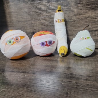 An orange, apple, banana, and pear wrapped to look like mummies with colorful googly eyes on a wood grain background