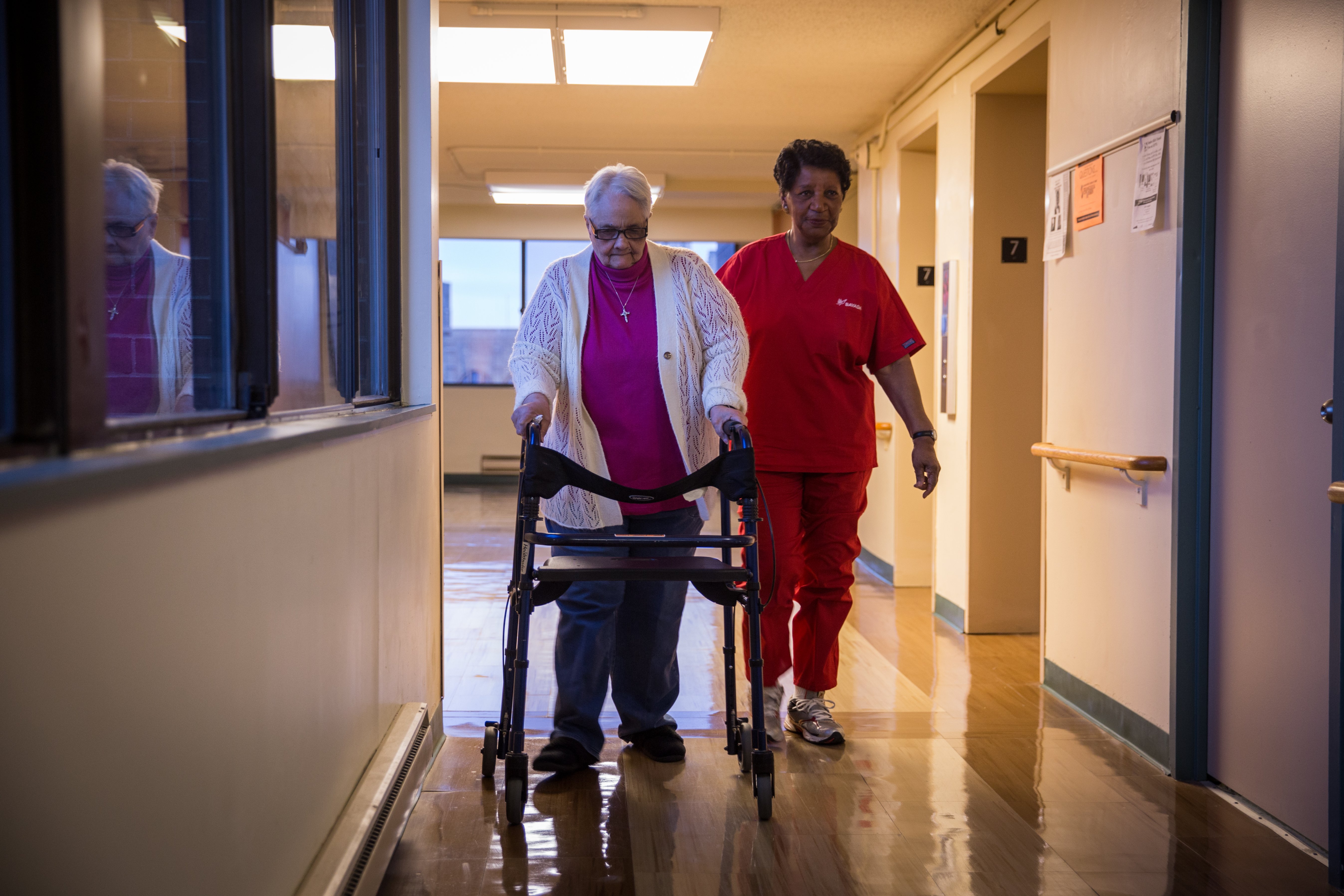 A home health aide leads her patient down a hallway.